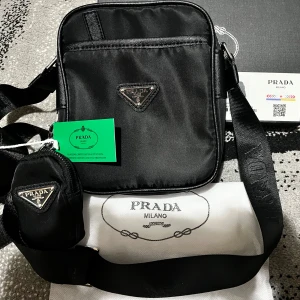 Svart axelväska med trekantig mönster  - Snygg svart axelväska  med klassisk trekantig logga framtill. Väskan har justerbar axelrem och en mindre avtagbar ficka. Tillverkad i ett glansigt syntetmaterial och levereras med dustbag och originalförpackning.