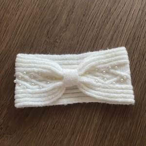 Vitt stickat diadem med pärlor - Snyggt vitt stickat diadem med ribbad struktur och dekorativa pärlor framtill. Diademet har en twistad knut i mitten och är perfekt för att hålla håret på plats under kyliga dagar. Lappen var bortklippt så jag sänker priset.