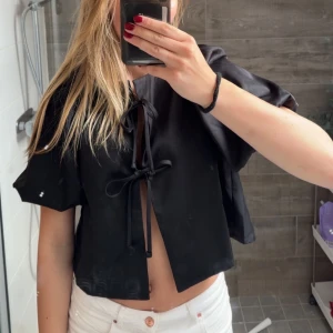 Svart blus med puffärmar och knytning - Svart blus med puffärm och två knytband framtill. Blusen är croppad och har en lös passform. På baksidan har det lite tråd spräckt upp (se sista bilden) men inget man märker💕💕 