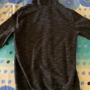 Mörkgrå hoodie med dragkedja från Champion - Säljer en mörkgrå zip-hoodie från Champion med stor logga i rött, vitt och blått på bröstet. Tröjan har huva med vita snören och två fickor framtill. Materialet är en blandning av bomull och polyester.