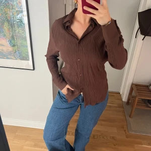 Brun skjorta från Bershka - Säljer en brun skjorta från Bershka med krage och knappar framtill. Skjortan har lång ärm och är tillverkad i ett lätt, strukturerat material som ger en snygg och avslappnad look. Perfekt att styla med jeans eller kjol.