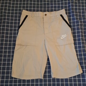 Nike shorts - Snygga beige shorts från Nike med svarta detaljer vid fickorna och klassisk Nike-logga på ena benet. Modellen är standard fit och har elastisk midja baktill för extra komfort. Perfekta för sommaren eller träning.