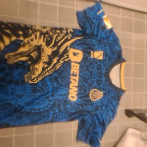 FC Porto blå fotbollströja med drakmotiv - Säljer en blå FC Porto fotbollströja från New Balance med ett stort gult drakmotiv på framsidan. Tröjan har korta ärmar, klubbmärke och sponsorloggor i gult. Baksidan har texten 'Super Bock' och ett litet FC Porto-märke i nacken. Storlek m men passar mig snyggt runt musklerna som L till XL