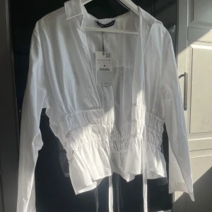 Vit omlottblus med knyt från Zara - Säljer en fräsch vit omlottblus från Zara med långa ärmar och knyt i midjan. Blusen har en v-ringad design och smockdetaljer som ger en snygg passform. Perfekt för en trendig och stilren look.