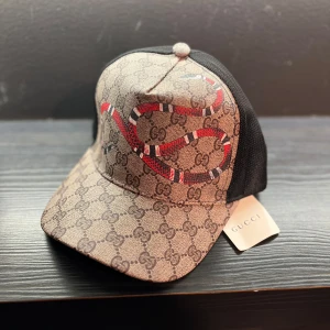 Keps med orm design - Keps men beige färg samt orm detaljer, kepsen är helt ny och säljes då den inte kommer till använding. Kepsen är trendig och passar perfekt till soliga dagarna. 