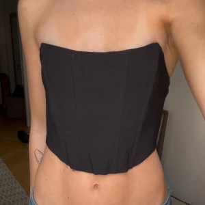 Svart tubtopp i korsettstil - Säljer en svart tubtopp med korsettkänsla. Toppen är croppad och har en stilren, figurnära passform utan axelband. Perfekt att styla med jeans eller kjol för en trendig look. Köpt från Sellpy men aldrig använd 💞