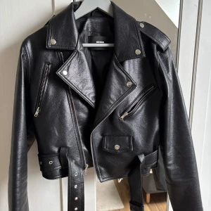Svart skinnjacka - Säljer en svart skinnjacka från Bik Bok med klassisk bikerstil. Jackan har silvriga dragkedjor, knappar, axelklaffar och bälte i midjan. Croppad modell med flera fickor och långa ärmar.