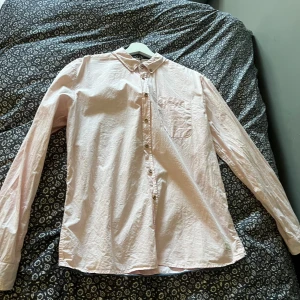 Ljusrosa skjorta med knappar - Säljer en ljusrosa skjorta med klassisk krage och bröstficka. Skjortan har långa ärmar och knäppning framtill med bruna knappar. Perfekt för en stilren och avslappnad look.