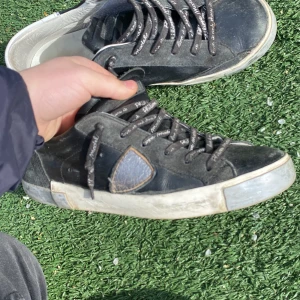 Svarta sneakers med grå detaljer - Säljer ett par svarta sneakers från Philippe Model med grå detaljer och vit sula. Skorna har snörning och en ikonisk sköld på sidan. Perfekta för dig som gillar en sportig och stilren look.