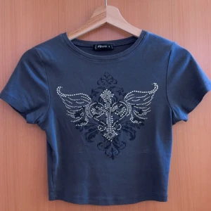 Mörkgrå croppad t-shirt med strassdetaljer från FB Sister - Snygg mörkgrå croppad t-shirt från FB Sister med korta ärmar. Framsidan har ett coolt tryck med vingar och kors i strass. Perfekt för dig som gillar edgy stil och vill sticka ut lite extra.