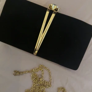 Svart kuvertväska med gulddetaljer och kedja - Snygg svart kuvertväska med guldfärgad kedja och dekorativt spänne med kristalldetalj. Väskan har en elegant form och passar perfekt när du vill lyfta din outfit med något extra.