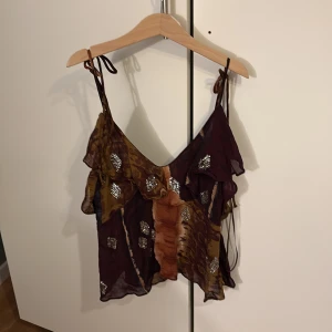 Brun och vinröd linnetopp från Zara - Säljer en unik linnetopp från Zara i bruna och vinröda nyanser med silvriga detaljer. Toppen har smala axelband som knyts och ett asymmetriskt, vågigt fall. Perfekt för dig som vill sticka ut.❤️