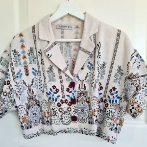 Mönstrad skjorta från Shenaz - Unik skjorta från Shenaz i beige med färgglatt grafiskt och blommigt mönster i blått, brunt och svart. Skjortan har korta ärmar, krage och tre bruna knappar framtill. Tillverkad i ett linneliknande material och har en avslappnad passform.