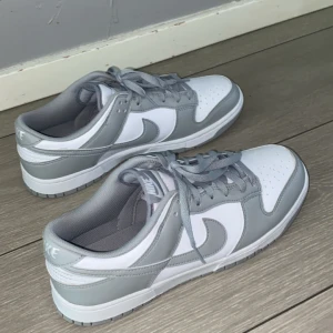 Nike Dunk Low i vitt och grått - Säljer ett par Nike Dunk Low sneakers i vitt och ljusgrått. Skorna har klassisk låg siluett, snörning och rund tå. Perfekta för dig som gillar stilrena och trendiga sneakers med ikonisk Nike-logga på sidan och tungan. Priset kan sänkas vid snabb affär köpta för 1400kr helt nya oanvända. säljer pga för stora köpta på JD Sports 