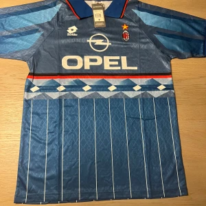 AC Milan retro bortatröja Baggio 18 - Klassisk blå AC Milan bortatröja från Lotto med OPEL sponsortryck och Baggio 18 på ryggen. Tröjan har vita och ljusblå ränder, röda detaljer vid kragen och klubbmärke på bröstet. Perfekt för samlare eller fans av italiensk fotboll.