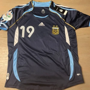 Argentina fotbollströja Messi #19 - Säljer en mörkblå Argentina fotbollströja från Adidas med Messi och nummer 19 på ryggen. Tröjan har korta ärmar, ljusblå och vita detaljer samt guldiga linjer. Perfekt för dig som älskar fotboll och vill stötta Argentina!