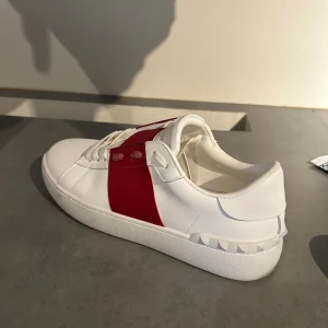 VO vit röda f4k3 - Helt nya aldrig använda! Snygga vita sneakers med en bred röd rem över ovansidan. Skorna har en rund tå och dekorativa nitar längs sulan. Perfekta för en stilren look. Säljer pga stor storlek 