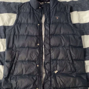 Mörkblå dunväst från Gant - Storlek 13-14år. Har ingen användning för den nåmer Mörkblå dunväst från Gant med broderad logotyp på bröstet. Västen har ståkrage, dragkedja och knappar framtill samt två fickor. Insidan är randig.