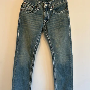 Blå jeansbyxor från True Religion med vita sömmar - Säljer ett par blå jeans från True Religion med tydliga vita kontrastsömmar och snygga detaljer på bakfickorna. Klassisk femficksmodell med knappgylf och dekorativa knappar. Perfekta för dig som gillar street och statement-jeans.💕{Längd:103,5 cm, Innerbenslängd: 78,5cm}