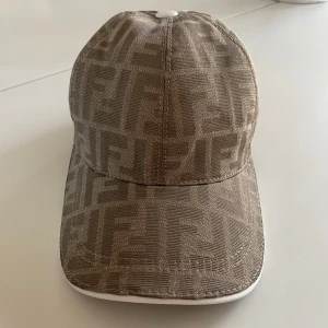 Beige keps från Fendi med monogram - Säljer en beige keps från Fendi med klassiskt FF-monogram över hela kepsen. Justerbart band bak i vitt läder med gulddetalj. Kepsen är tillverkad i 100% bomull och har en böjd skärm.