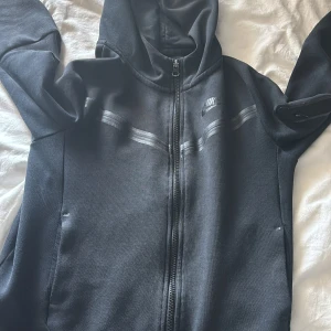 Svart hoodiejacka från Nike med dragkedja - Säljer en svart hoodiejacka från Nike med hel dragkedja framtill och huva. Jackan har diskreta svarta detaljer och fickor med dragkedja på ärmarna. Perfekt för en sportig stil och enkel att matcha.
