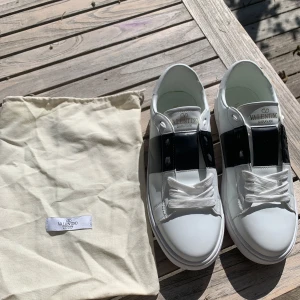 Valentino Garavani Open sneakers i vitt och svart - Använda 2 gånger men känner att dem är lite för små så säljer pga det! Så inprincip helt nya! Säljer ett par snygga Valentino Garavani Open sneakers i vitt skinn med bred svart rem över mitten. Skorna har vita snören och diskret Valentino-logga i guld på plösen och innersulan. Medföljer originalpåse. 