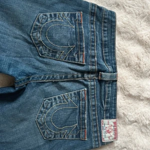 True Religion blå bootcut jeans - Säljer ett par blå jeans från True Religion med slitna detaljer på benen. Jeansen har bootcut-modell, orange sömmar och klassisk logga på bakfickan. 