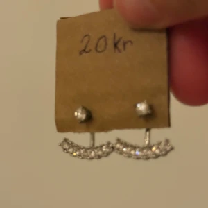 Glittriga örhängen i silverfärg - Säljer ett par snygga örhängen i silverfärg med glittrande stenar. Örhängena har en unik form där en rad med små stenar hänger under örsnibben och ger en cool effekt. Perfekta för dig som vill ha något extra till din look.