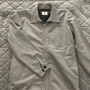 Grå overshirt från SAMS XS - Snygg grå overshirt från SAMS i storlek XS. Jackan har klassisk krage, dragkedja framtill, en bröstficka och knappar vid ärmsluten. Perfekt för lager-på-lager och enkel att matcha med det mesta.