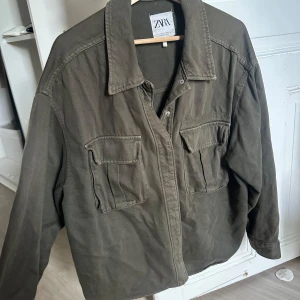 Olivgrön jeansjacka från Zara - Snygg olivgrön oversized jeansjacka från Zara med klassisk krage och två stora bröstfickor med lock. Jackan är i bomull och har en avslappnad passform, perfekt att slänga över en hoodie eller t-shirt. Enkel knäppning framtill och långa ärmar.
