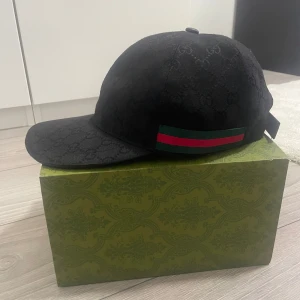 Svart keps från Gucci - Snygg svart keps från Gucci med diskret mönster och klassiska röd-gröna ränder på sidan. Perfekt för en stilren look.