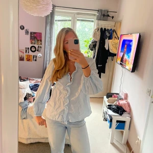 Ljusblå volangskjorta i bomull - Supersöt ljusblå skjorta med volang längs v-ringningen och framtill. Skjortan har långa ärmar med resår vid handlederna och är gjord i ett tunt bomullstyg. Perfekt för dig som gillar en romantisk och chill stil.