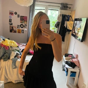 Svart axelbandslös kortklänning - Säljer en svart axelbandslös kortklänning med volang nertill och snygg rynkdetalj längs sidan. Klänningen har en tight passform upptill och är tillverkad i ett mjukt, stretchigt material som känns bekvämt mot huden.