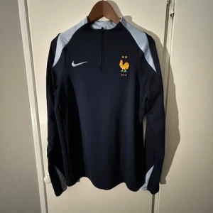 Frankrike Nike långärmad fotbollströja - Mörkblå långärmad fotbollströja från Nike med Frankrikes landslagsemblem på bröstet och ljusblå detaljer vid axlar och ärmslut. Tröjan är i Dri-Fit-material och har en halv dragkedja framtill. Slim fit-modell, perfekt för träning eller match.