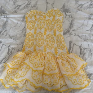 Gul och vit axelbandslös kortklänning - Superfin axelbandslös kortklänning i vit och gul med broderade detaljer och volang nedtill. Klänningen har ett smockat liv och ett skirt, broderat tyg med blommönster på kjolen. Perfekt för sommarens alla tillfällen! Köpta utomlands ❤️