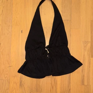 Svart halterneck topp från Nelly - Supersnygg svart halterneck topp från Nelly med volangkant nertill och snörning framtill. Toppen har en croppad passform och är perfekt för att skapa en trendig look. Tillverkad i ett mjukt, lätt material som känns skönt mot huden.