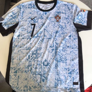 Portugal Ronaldo #7 fotbollströja Nike - Portugal fotbollströja med Ronaldo och nummer 7. Tröjan är kortärmad med vit bas och blått mönster, svarta detaljer på krage och ärmslut. Nike-logga och Portugals landslagsemblem på bröstet. Tillverkad i lätt och ventilerande material, perfekt för match eller träning. Klubbloggan på bröstet är lite skadat