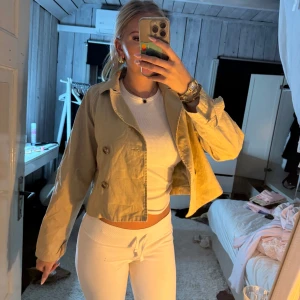 Beige trenchcoat - Säljer en beige trench coat med dubbelknäppning och klassisk krage. Jackan har långa ärmar och är tillverkad i ett bomullsliknande material. Perfekt för dig som gillar en clean och stilren look med lite edge.
