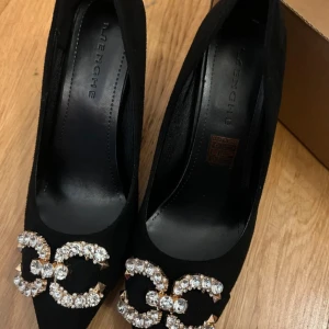 Svarta pumps med strassdetalj - Snygga svarta pumps med spetsig tå och hög klack. Skorna är i mockaliknande material och har en glittrig dekoration framtill med stora strasstenar och guldfärgade nitar. Perfekta för dig som vill sticka ut med en elegant och trendig look. Helt oanvända och bekväma.