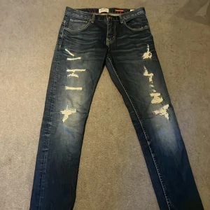 Crocker 310 Slim slitna jeans 30/34 - Mörkblå slim fit jeans från Crocker, modell 310 Slim. Snygga slitningar och distressed detaljer framtill för en cool look. Klassisk femficksmodell med dragkedja och knapp. Perfekta för dig som gillar en smalare passform