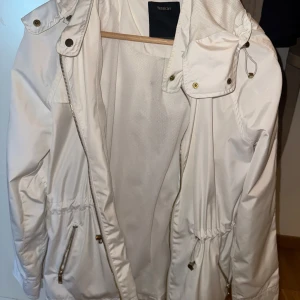 Vit vind/regnjacka från Yessica strl 36 - Snygg vit vind/regnjacka från Yessica med huva, dragkedja och snörning i midjan. Jackan har meshfoder, guldiga detaljer och praktiska fickor med dragkedja. Perfekt för dig som gillar stilren och sportig look. Materialet är polyester.