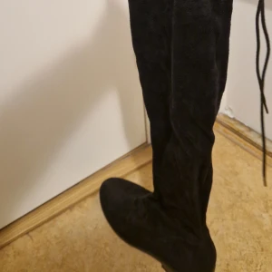 Svarta overknee mockastövlar - Snygga svarta overknee-stövlar i mjuk mocka med snörning upptill. De har platt sula och rund tå, perfekt för dig som vill ha en stilren och trendig look. Passar till både kjol och jeans för en cool vibe.