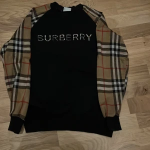 Burberry svart tröja med rutiga ärmar - Svart tröja från Burberry med klassiska rutiga ärmar i beige, brunt, vitt och rött. Stor Burberry-logga framtill. Tröjan har långärmad passform och ribbade muddar. Perfekt för dig som gillar ikoniska designer och vill sticka ut med stil.