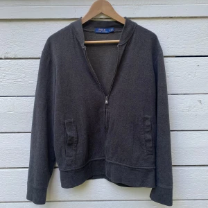 Grå zip hoodie från Polo Ralph Lauren - Mörkgrå hoodie från Polo Ralph Lauren med hel dragkedja framtill och två fickor. Klassisk och enkel design med lång ärm och mjukt bomullsmaterial. Perfekt för en chill och stilren look.