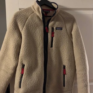 Beige teddyjacka från Patagonia M - Säljer en beige teddyjacka från Patagonia i storlek M. Jackan har mysigt teddyfoder, hög krage och tre fickor med dragkedja och röda detaljer. Perfekt för kyliga dagar och riktigt skön att ha på sig. Klassisk Patagonia-logga på bröstet. Finns ej längre att köpa
