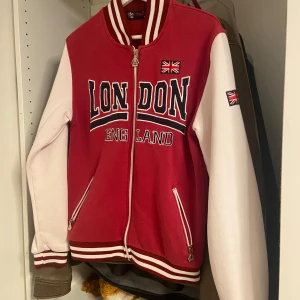 Jacka / Kofta - Oversized röd och vit jacka / kofta från G-Crown med broderad text 'London England' och brittiska flaggor. Jackan har dragkedja, randiga muddar och två fickor framtill. Perfekt för dig som vill sticka ut med en sportig och urban stil. Den är som en storlek M/L.