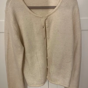 Beige stickad kofta med knappar - Mysig beige stickad kofta med rund halsringning och knäppning framtill. Långärmad och enkel design som passar perfekt till lager-på-lager. Koftan har en mjuk känsla och neutrala knappar som smälter in i plagget.