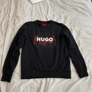 Svart sweatshirt från Hugo Boss XS - Svart sweatshirt från Hugo Boss med coolt HUGO-tryck och röda flames på bröstet. Tröjan har rund hals, långa ärmar och ribbade muddar. Tillverkad i mjuk bomull som är skön att ha på sig. Perfekt för dig som gillar streetstyle och vill sticka ut. Helt nytt skick säljer den för att den börjar bli liten har haft den i ungefär 3 månader köpt för 850 