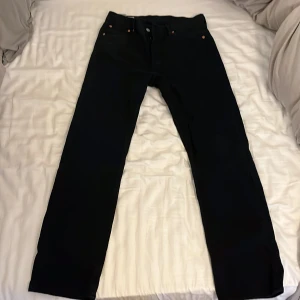 Svarta Levi's 501 jeans W30 L32 - Klassiska svarta Levi's 501 jeans med raka ben och knappgylf. Jeansen har fem fickor, snygga kopparfärgade nitar och den ikoniska läderpatchen bak i midjan. Tillverkade i slitstarkt jeanstyg, perfekta för en clean och tidlös look.