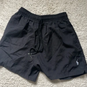 Svarta shorts från Polo Ralph Lauren - Svarta shorts från Polo Ralph Lauren med klassisk broderad logga i vitt på benet. De har elastisk midja med snörning för skön passform och är gjorda i mjuk bomull. Perfekta för chill dagar och sommarhäng.